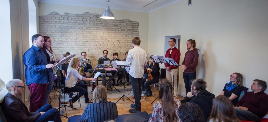 A man stood in the middle of a room conducting a small number of musicians in front of an audience. 