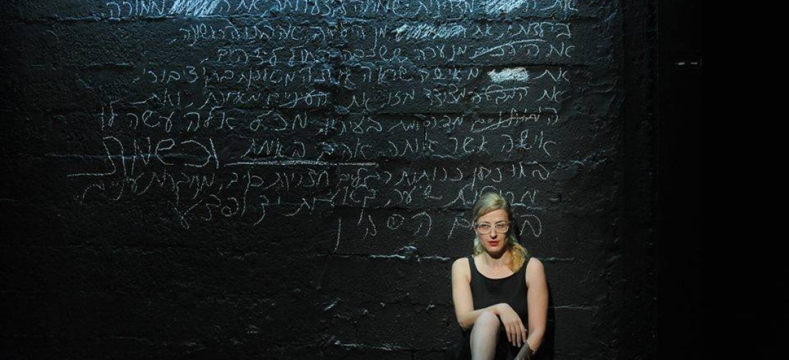A performer sitting in front of a black wall covered in white chalk writing. 