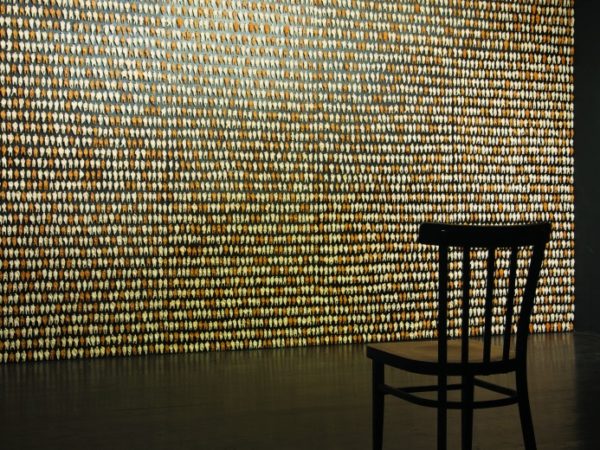A wooden chair placed in front of a wall filled with rows of very small models of people in either a white or terracotta colour.