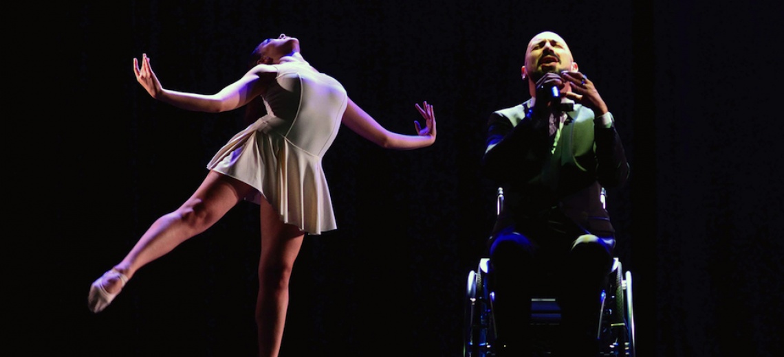 A man in a wheelchair holding a microphone and a woman in a ballet pose. 