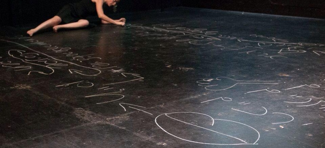 A performer lying on a black stage floor which is covered in chalk writing and pictures. 