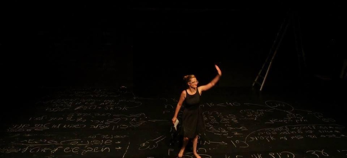 A performer standing waving on a black stage floor which is covered in chalk writing and pictures. 