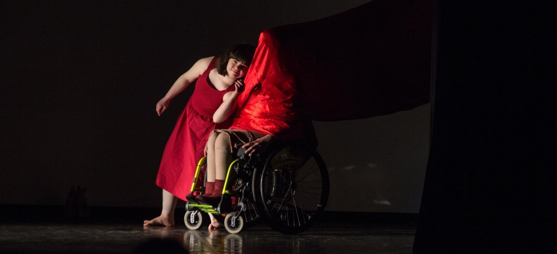 Two people on a dark stage. One is in a wheelchair with a red cloth draped over their head and body.
