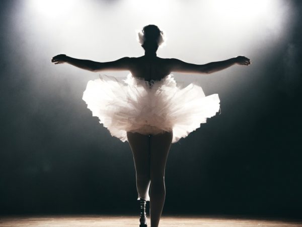 The back of a dancer with a prosthetic lower left limb, standing with arms outstretched in a ballet pose.