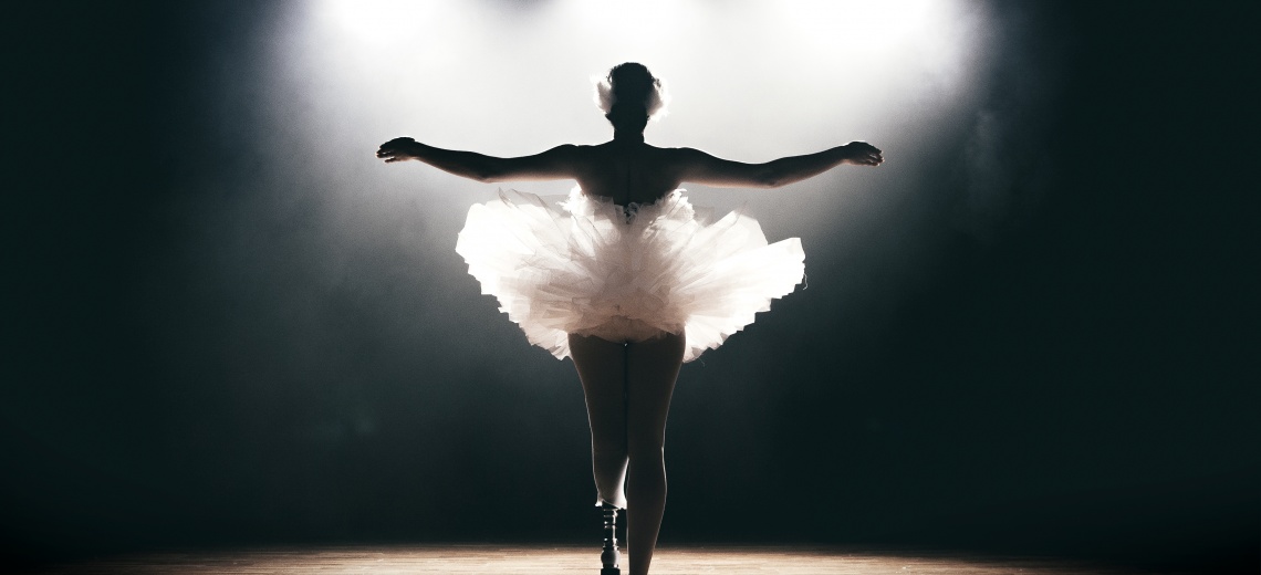 The back of a dancer with a prosthetic lower left limb, standing with arms outstretched in a ballet pose.