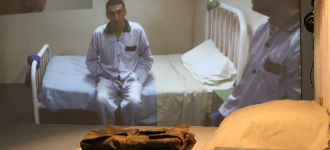 Two men wearing pyjamas, one of whom is sitting on the edge of a white metal framed bed, the other is in partial shot, in the foreground of the photograph.
