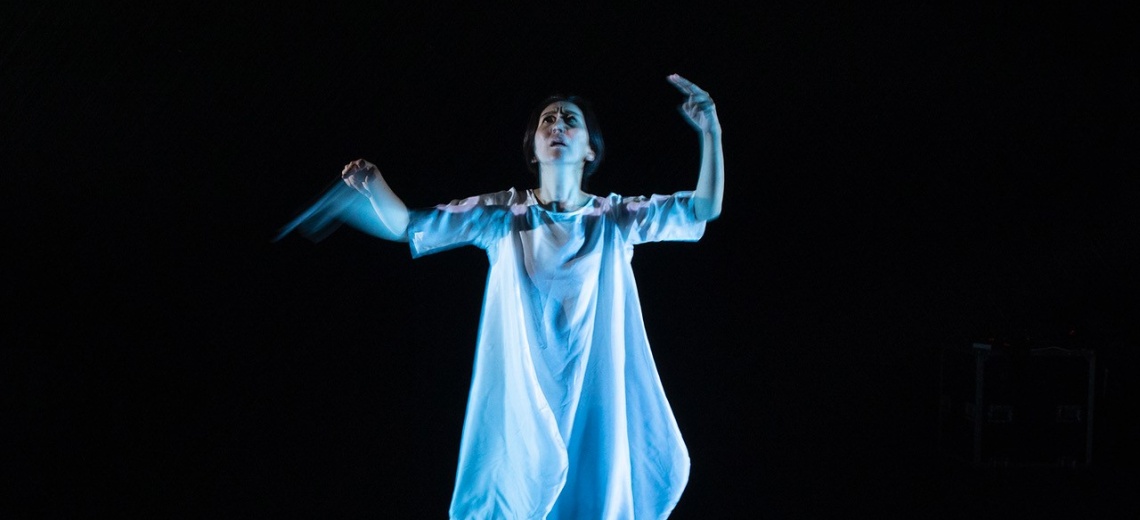  A woman on a dark stage looking upwards and with raised arms. 