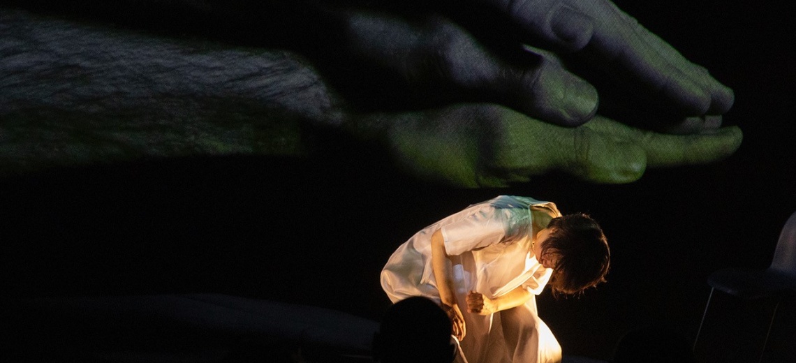  A woman on a dark stage bent over. There is a large projection of hand on hand in the background. 