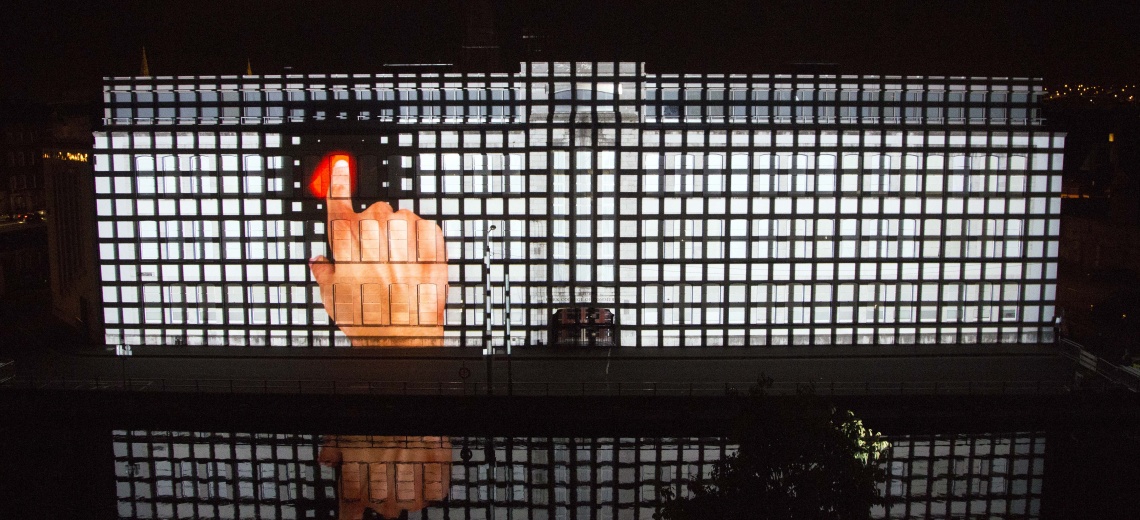 A light projection onto a building of a large hand pressing a red switch. 