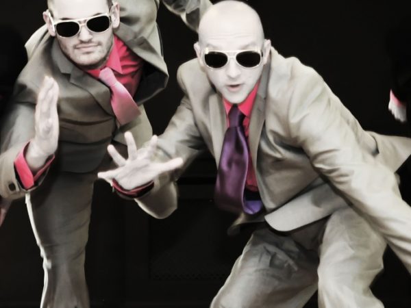 Four performers posing on stage, wearing suits and dark glasses