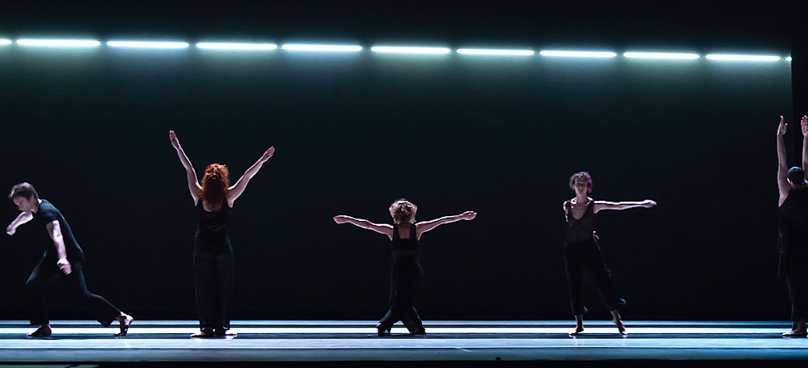 Five dancers on a dark stage in different poses with outstretched arms. 