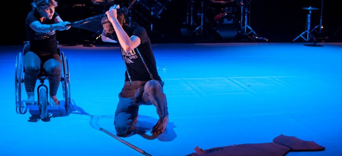 Two people on a stage. One is in a wheelchair and the other is kneeling nearby. 