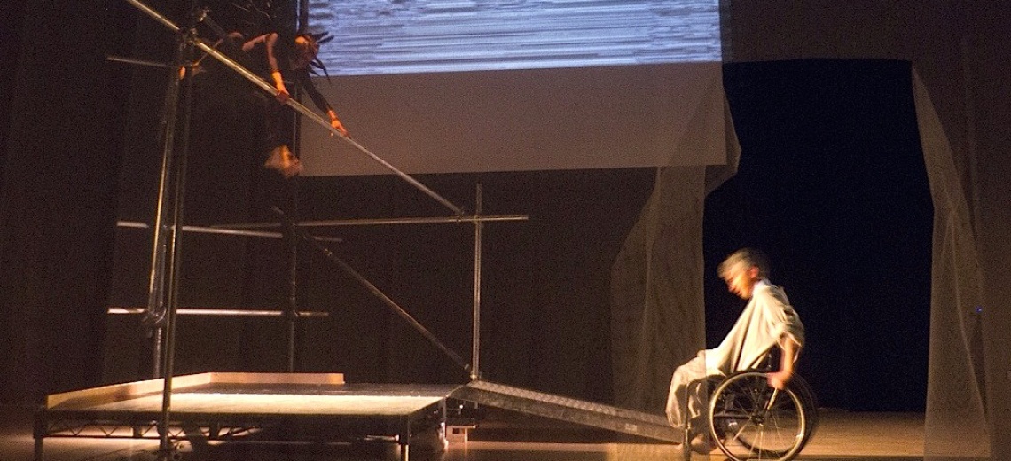 Two performers on a stage. One is on a scaffold platform, and the other is in a wheelchair at the bottom of a ramp. 