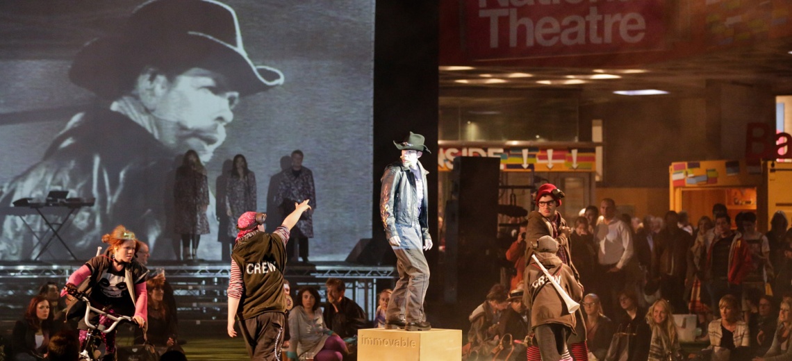 A group of performers in a busy open space, one is on a plinth wearing a cowboy hat. 