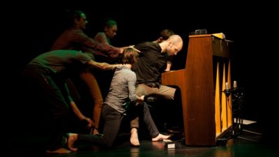 Five people appear to be pulling a person away from their seat at a piano