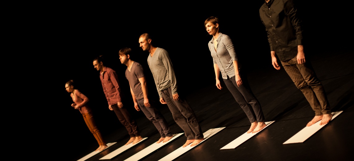 six people standing barefoot, on white boards which are placed at regular intervals on a black floor