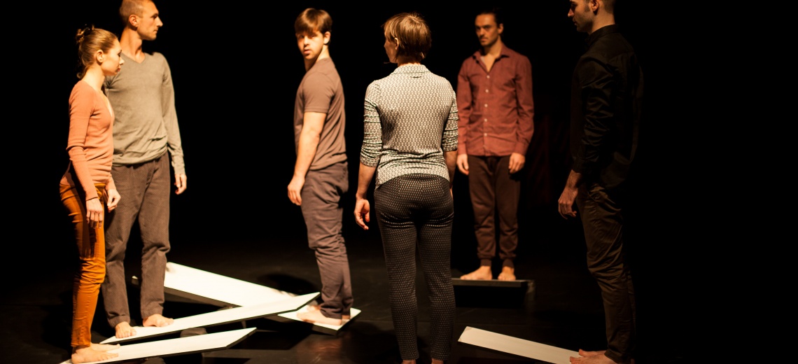 Six people standing barefoot on a stage, five people are stood on see-saw like on planks