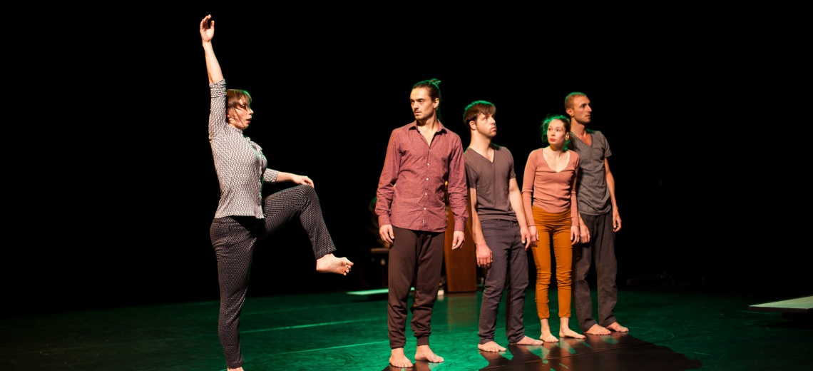 Five people are standing barefoot on a stage. Four are in a row and a fifth is standing on one leg with their hand in the air. 