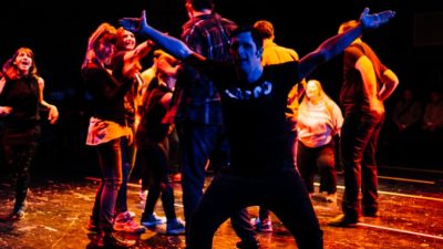 A group of performers dancing on a stage. One performer is standing with his arms and legs outstretched.