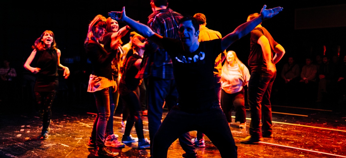 A group of performers dancing on a stage. One performer is standing with his arms and legs outstretched. 