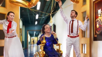 Three performers in a long corridor, one performer is in a power assisted chair.