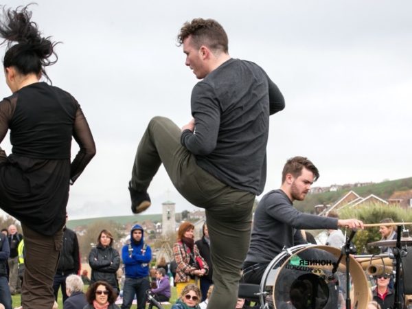 A man and a woman dancing in the open air in front of a small crowd. There is a man playing a drum kit in the background.