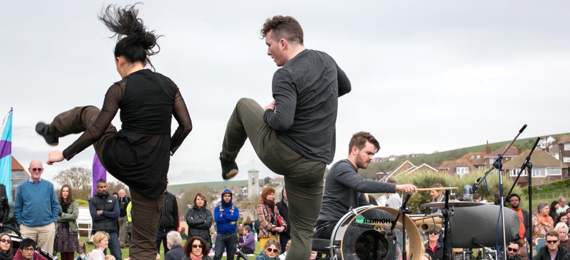 A man and a woman dancing in the open air in front of a small crowd. There is a man playing a drum kit in the background. 