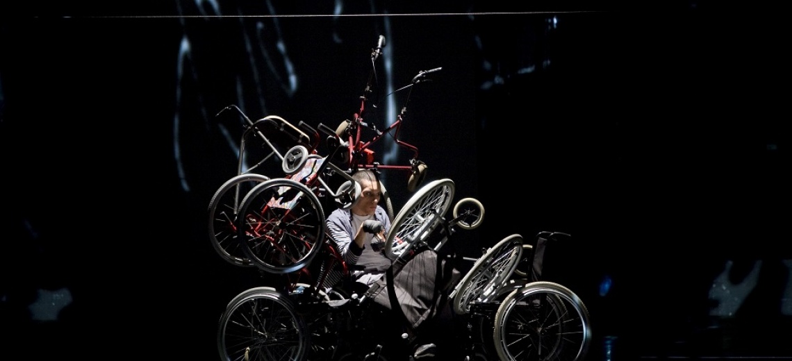 A man on a stage who is positioned under a tangle of wheels and metal frames. 