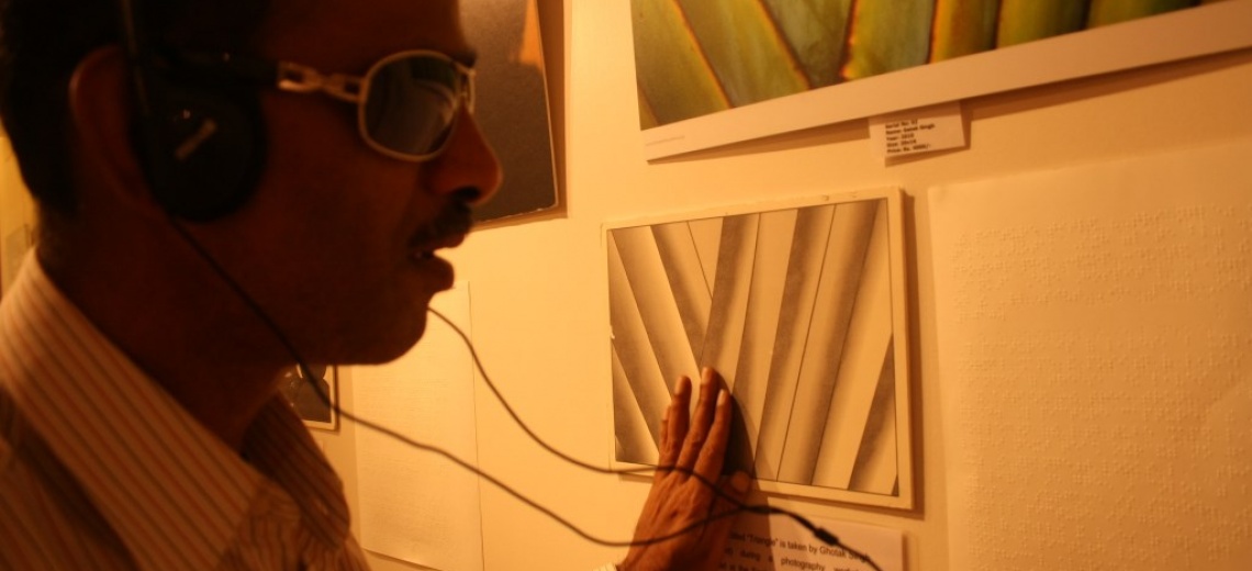 Close up of the face of a man wearing dark glasses and headphones, his hand is feeling a picture on the wall in front of him