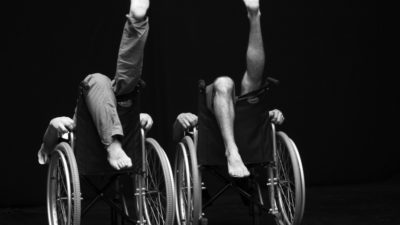 Two people sitting back to front in wheelchairs with their legs in the air.