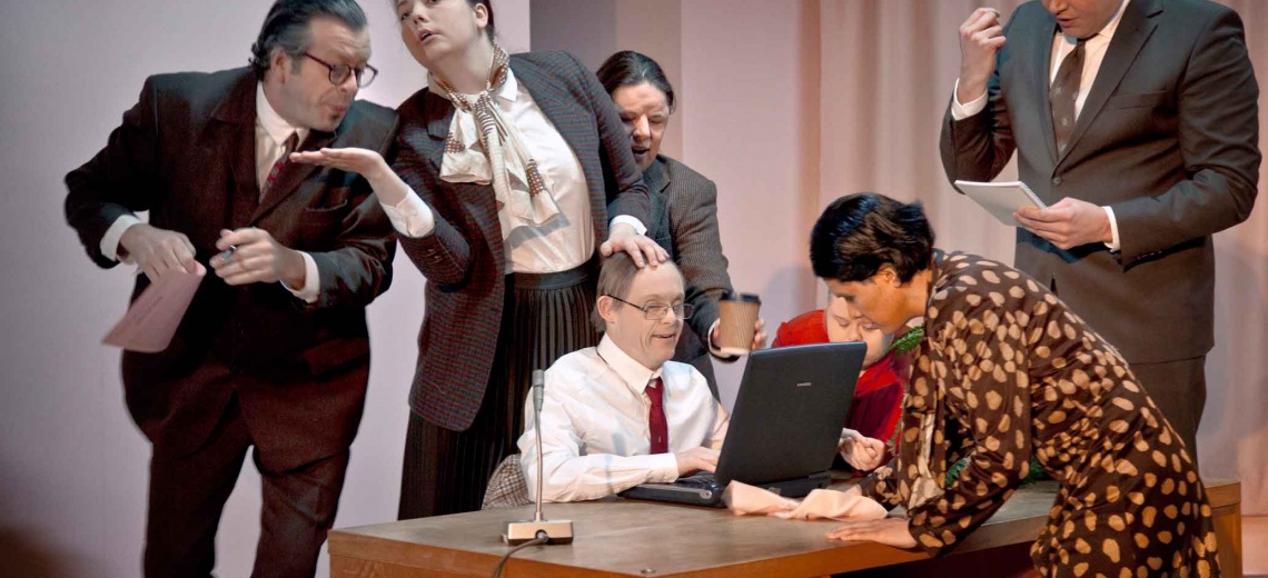 Seven performers on a stage, all crowded around a desk.