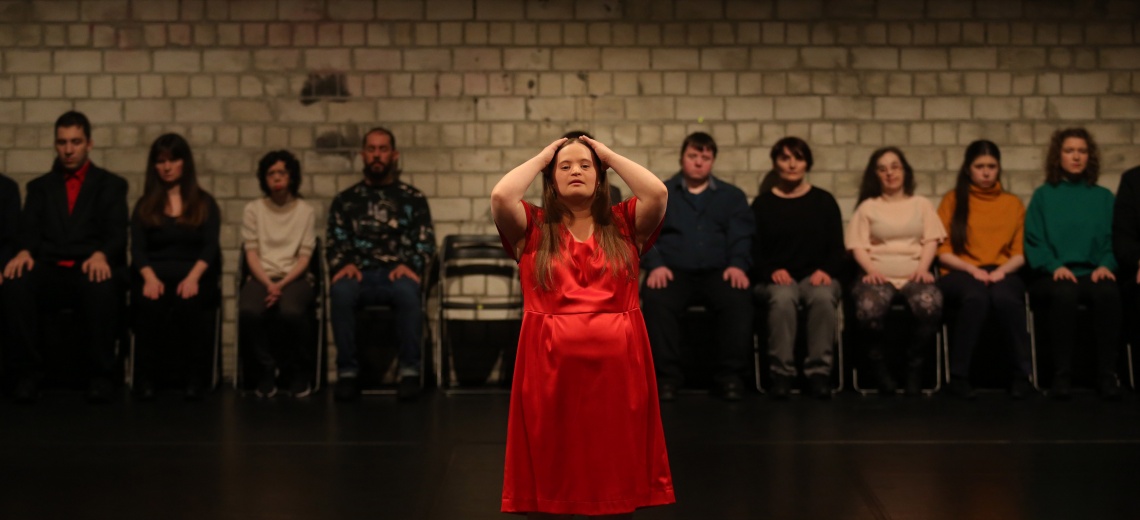 Performer in the centre of the scene, hands raised on her head. A group of people are sitting on chairs in the background. 