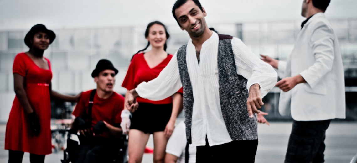 A man stood in a pose at the front of the scene, with three people wearing red in the background looking towards the camera. A fifth person is facing away form the camera to the right of the picture. 