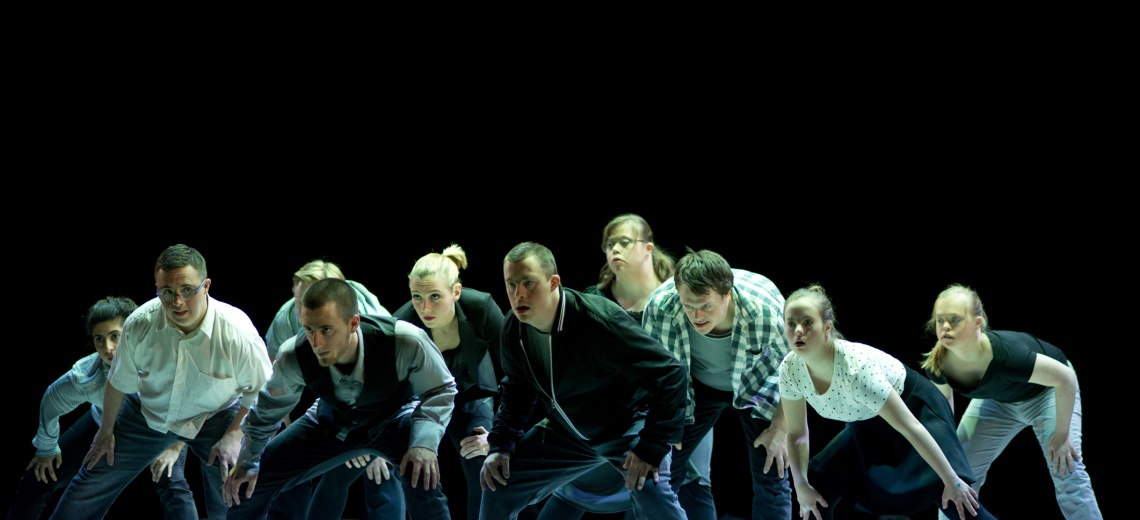 A group of 10 performers on a stage all leaning forwards in the same direction.