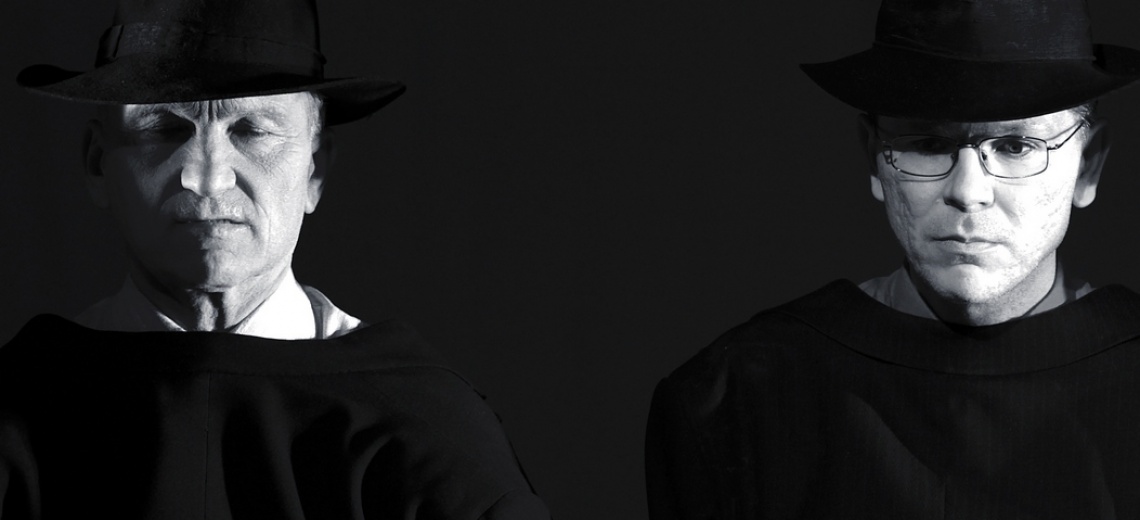 Two men wearing trilby hats, their faces highlighted against a dark background. 