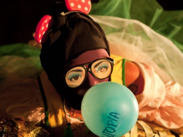 A person in a balaclava and large glasses blowing a balloon.