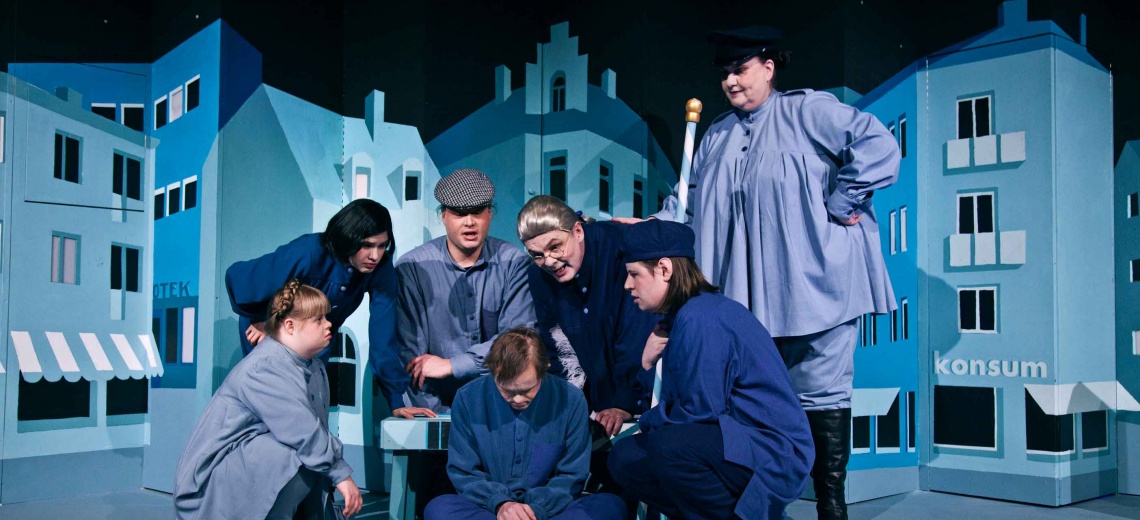 Seven performers huddled together on a stage all dressed in blue. 