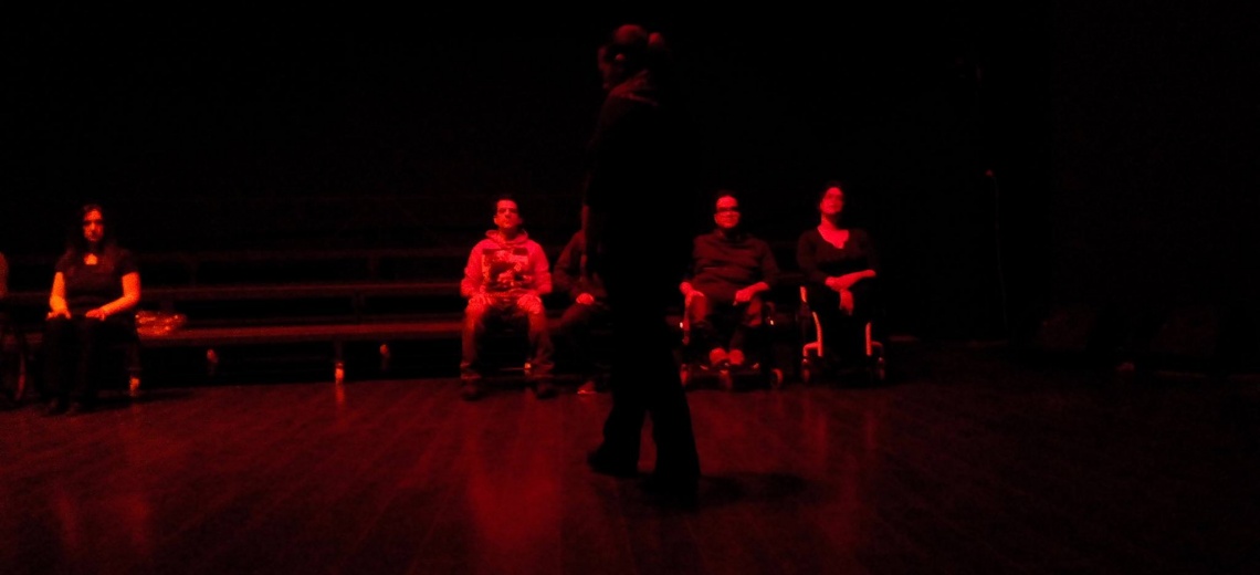 5 seated performers, whilst one paces, cast in red light