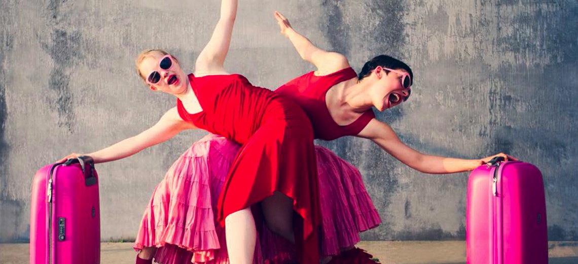 Two dancers all in red crossing each other and holding on to pink suitcases