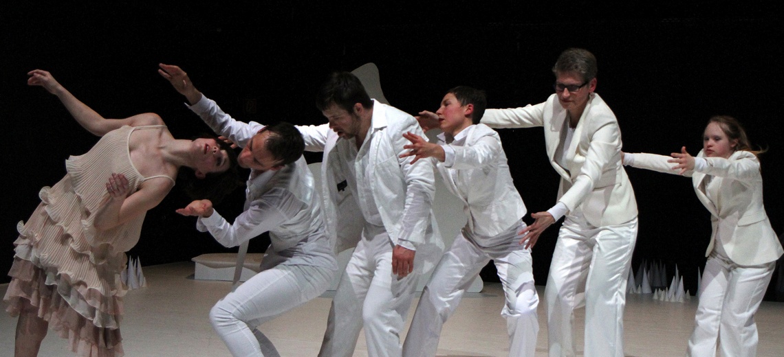 5 dancers in all white suits stand behind a woman in a beige dress who is leaning backwards