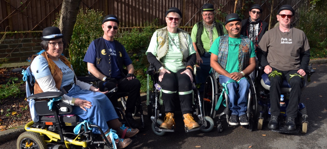 Seven performers in wheelchairs, wearing bowler hats, in a garden setting. 
