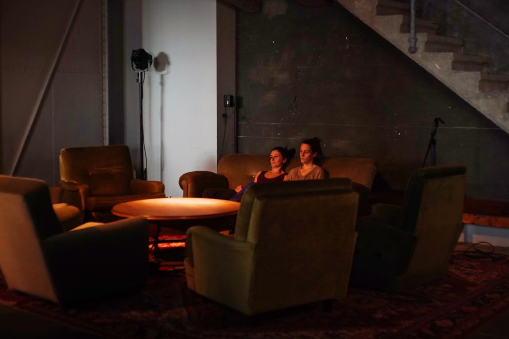 Two female dancers sit back on a dimly lit sofa, their eyes looking ahead, the scene with a rouge glow
