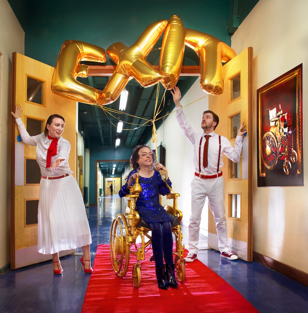 Liz Carr in a golden wheelchair with balloons spelling exit above her