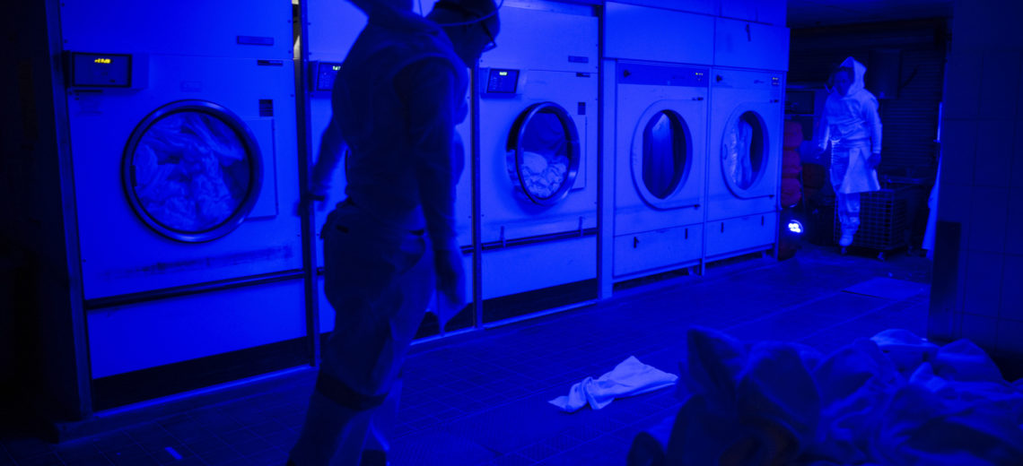 Two performers suspended from the ceiling in a launderette setting. 