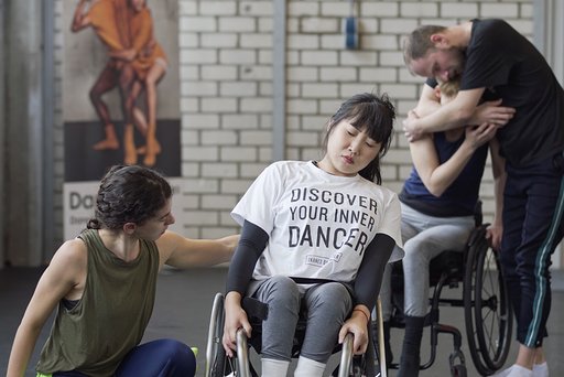 A dancer in a wheelchair closes her eyes and sways to one side with a dancer on the floor reaching her arm towards her
