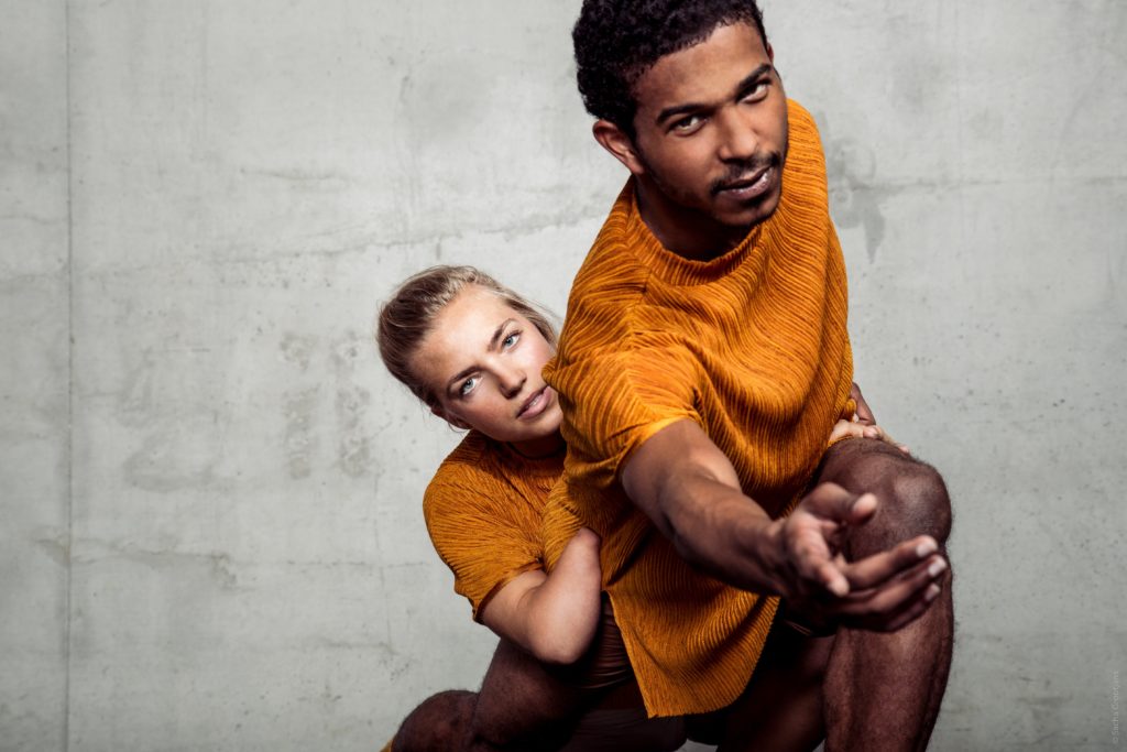 A male and female dancer dressed in matching orange tops reach out towards you.