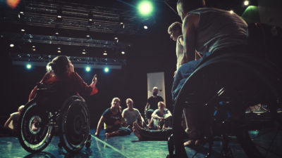 An artist in a wheelchair addresses a group of artists gathered round to listen