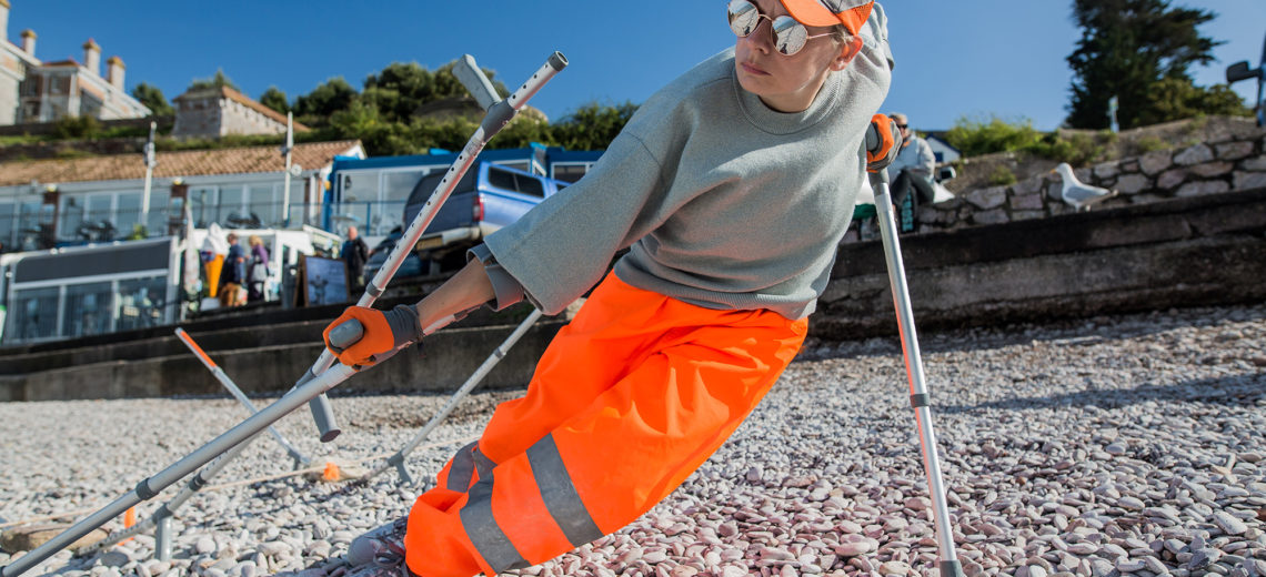 A woman using crutches stands on a pebble beach