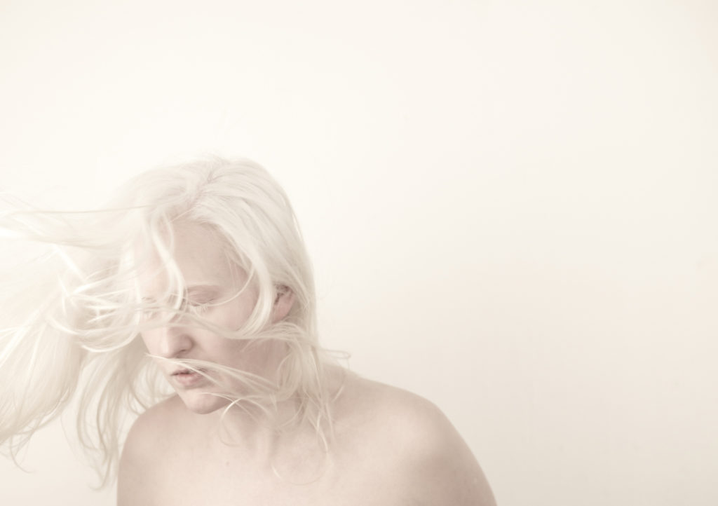 An artist with very white hair and pale skin looks away, pictured against a stark white background