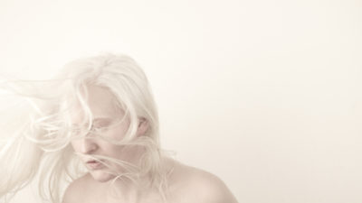 An artist with very white hair and pale skin looks away, pictured against a stark white background